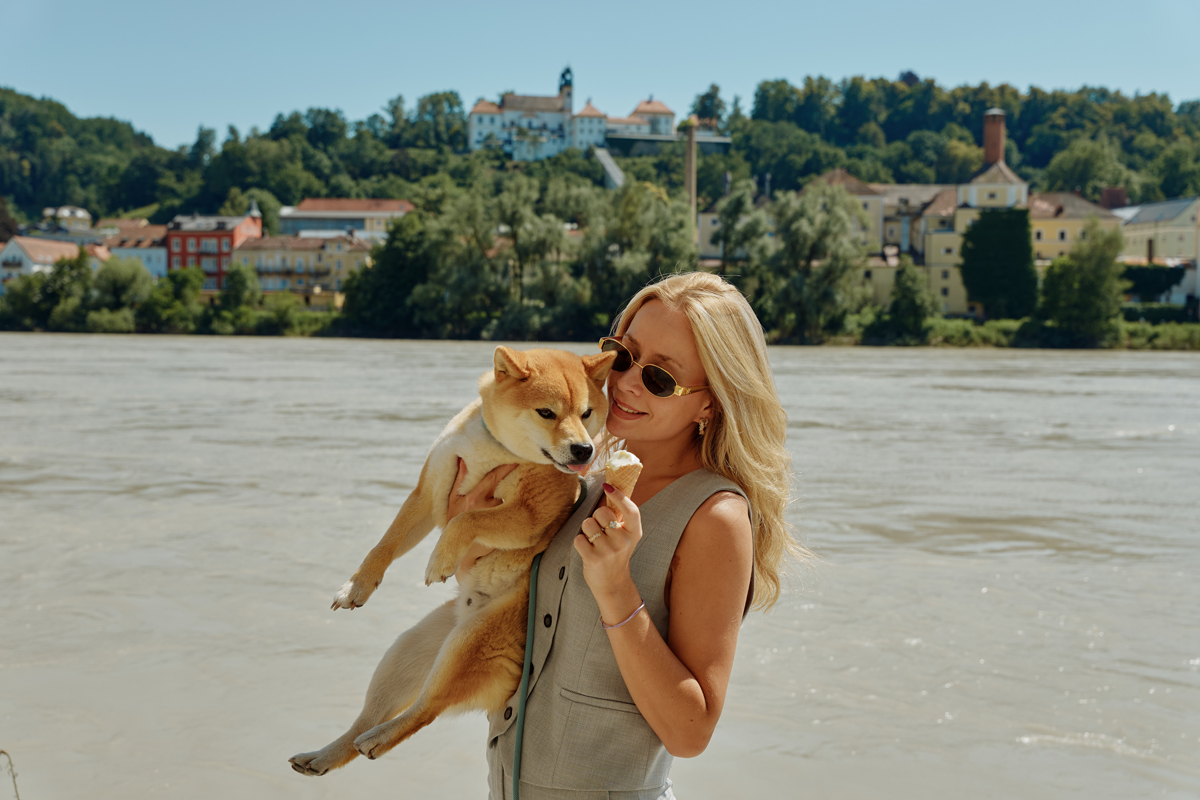 Eine Frau mit Sonnenbrille hält einen Shiba Inu und bietet ihm spielerisch eine Eistüte an, während sie an einem Fluss steht. Im Hintergrund ist eine malerische Aussicht auf Gebäude und einen bewaldeten Hügel unter einem klaren blauen Himmel zu sehen.