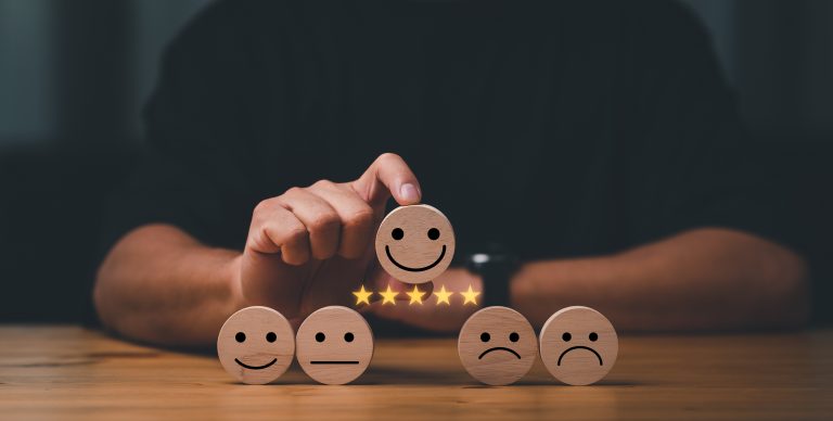 Eine Person wählt einen Holzblock mit einem lächelnden Gesicht und fünf Sternen darunter aus, umgeben von Blöcken mit neutralen und traurigen Gesichtern, die verschiedene Stufen der Zufriedenheit oder des Kundenfeedbacks darstellen.