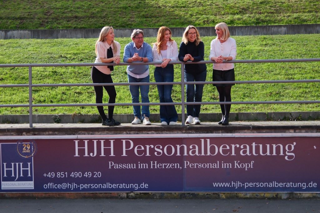 Fünf Personen stehen und unterhalten sich an einem Metallgeländer vor einem Schild der HJH Personalberatung, mit grünem Gras im Hintergrund. Die Gruppe wirkt entspannt und freundlich, einander zugewandt, im Freien an einem sonnigen Tag.