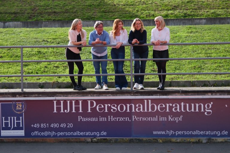 Fünf Personen stehen und unterhalten sich an einem Metallgeländer vor einem Schild der HJH Personalberatung, mit grünem Gras im Hintergrund. Die Gruppe wirkt entspannt und freundlich, einander zugewandt, im Freien an einem sonnigen Tag.