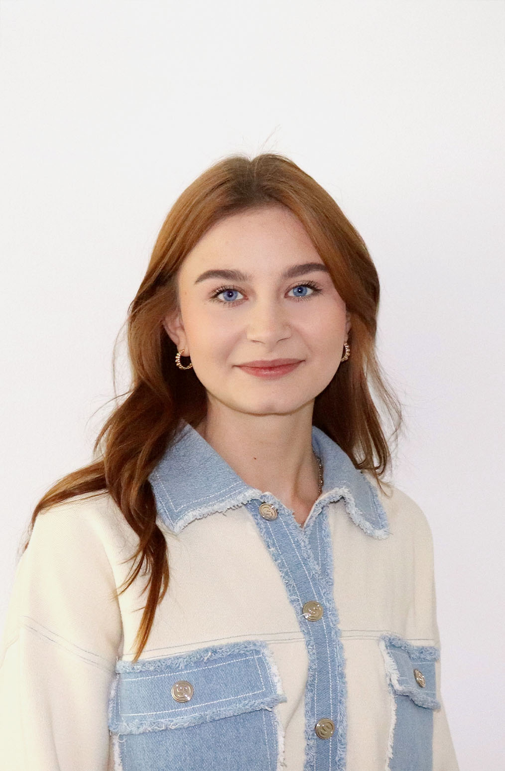Eine junge Frau mit langen braunen Haaren trägt ein cremefarbenes und jeansfarbenes Button-up-Hemd, goldene Ohrringe und lächelt leicht vor einem schlichten weißen Hintergrund.
