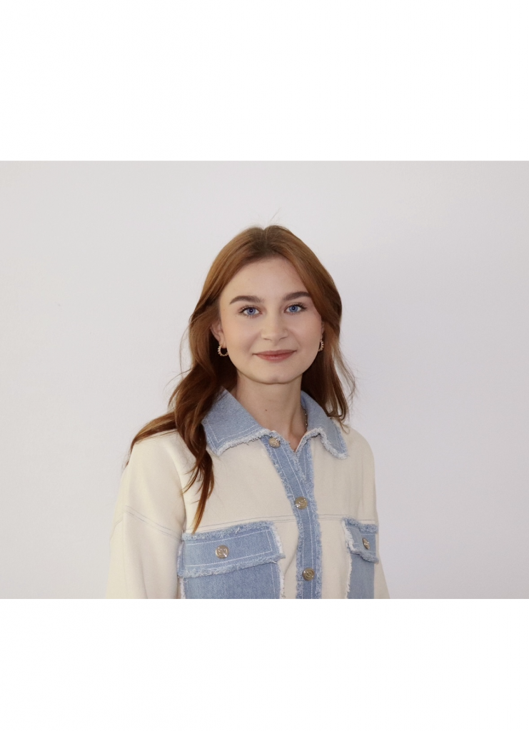 Eine junge Frau mit langem, hellbraunem Haar und neutralem Gesichtsausdruck steht vor einem schlichten weißen Hintergrund und trägt ein helles Hemd mit Jeans-Akzenten.