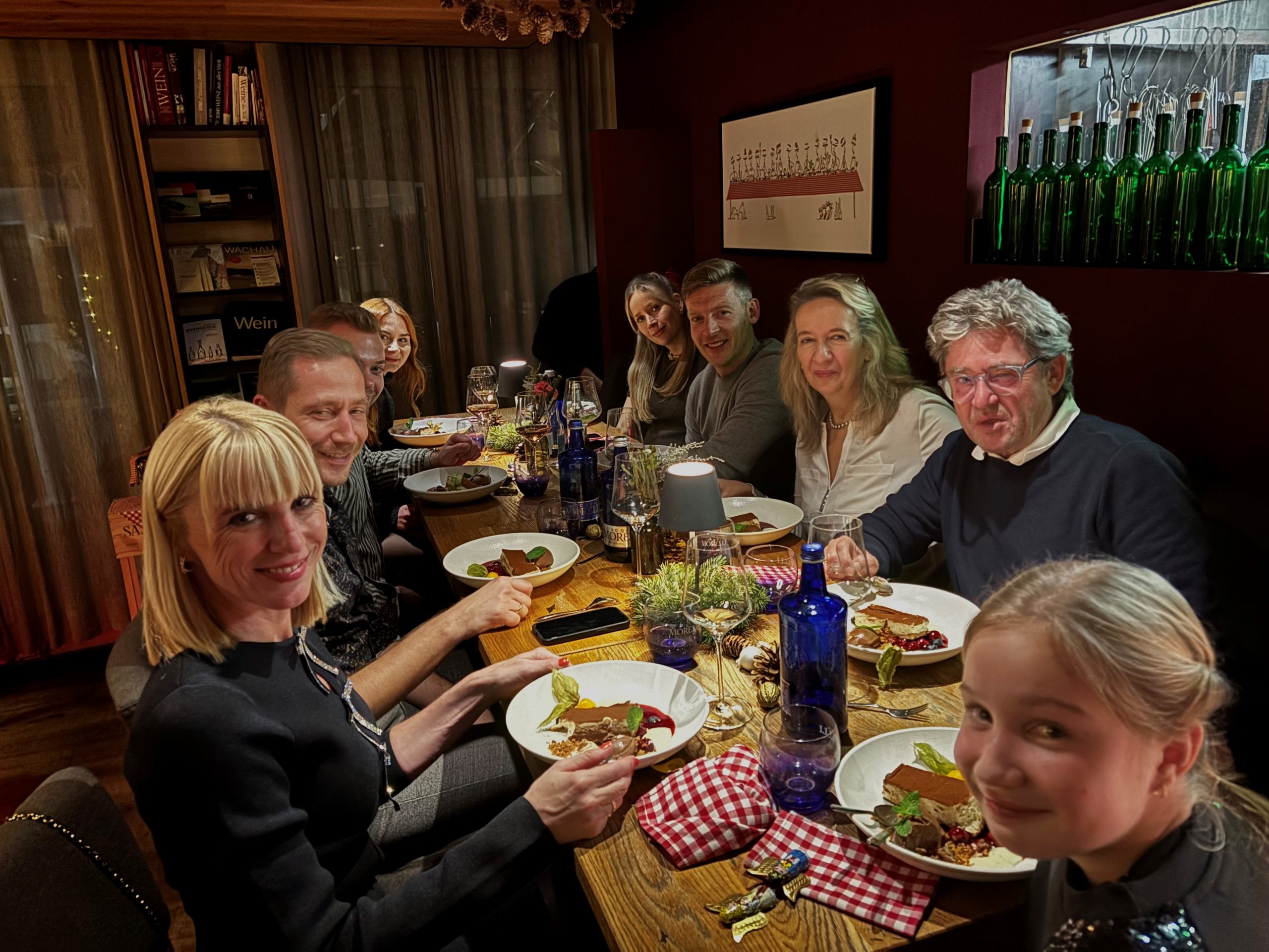 Eine Gruppe von neun Personen sitzt um einen warm beleuchteten Esstisch, teilt eine Mahlzeit und lächelt in die Kamera in einem gemütlichen, dekorierten Raum mit Regalen und Flaschen im Hintergrund.